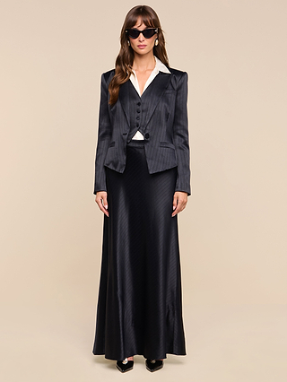 A woman stands facing forward against a plain beige background, wearing sunglasses, a white-collared shirt, the Jianna Vest—a black pinstripe blazer with a minimalist silhouette—and a long black satin skirt.