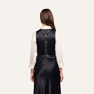 A woman stands with her back to the camera, wearing the Jianna Vest—a black pinstripe vest dress layered over a white long-sleeve shirt. This minimalist suiting style features black straps and silver buckles across the back.