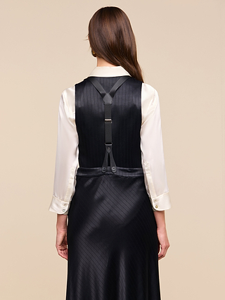 Wearing the Jianna Vest over a cream satin blouse, a woman with long brown hair, seen from behind, creates a striking minimalist silhouette. The silky black pinstripe vest features black leather suspenders attached to it for an effortlessly modern look.
