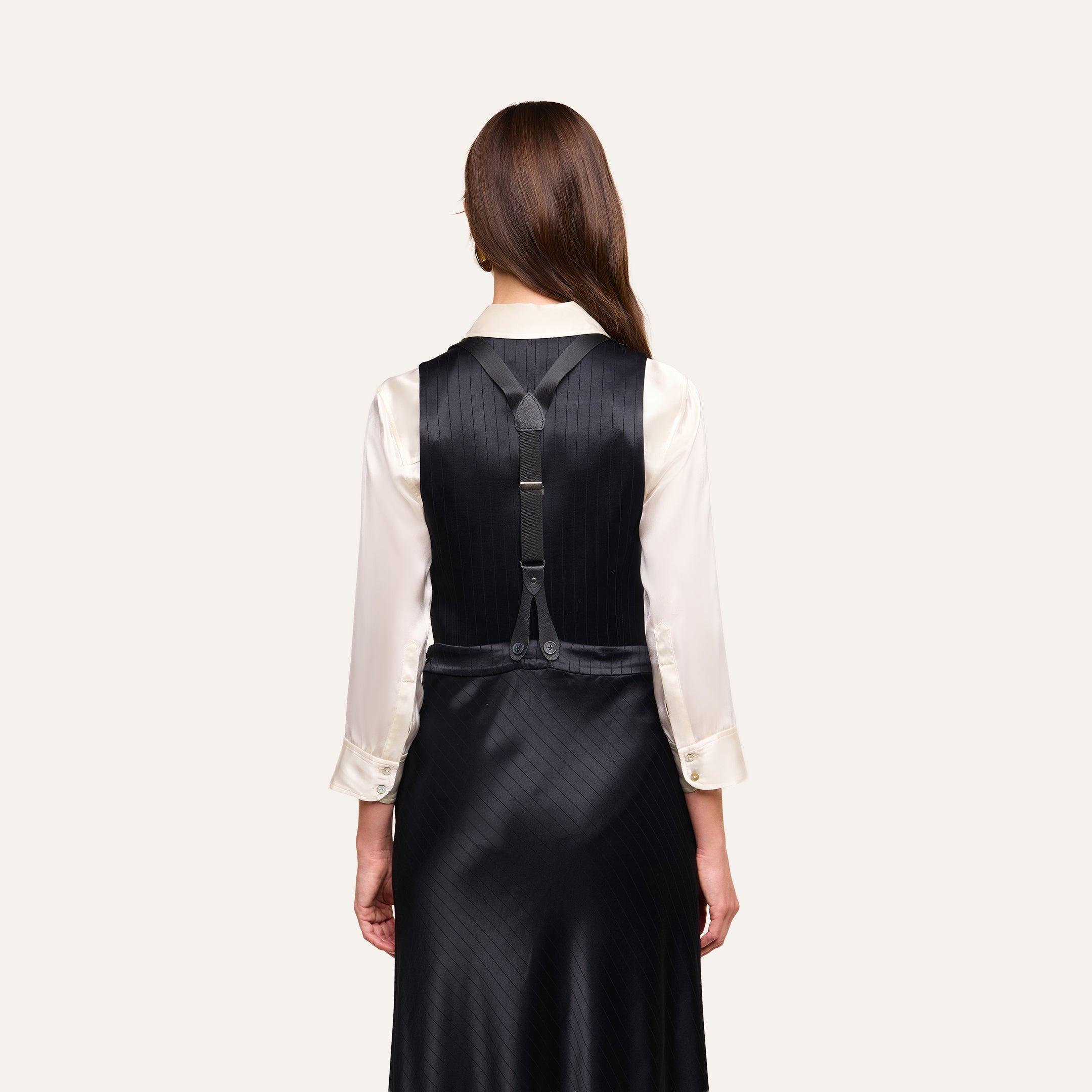A woman stands with her back to the camera, wearing the Jianna Vest—a black pinstripe vest dress layered over a white long-sleeve shirt. This minimalist suiting style features black straps and silver buckles across the back.