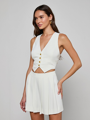 A woman with long brown hair and gold hoop earrings poses against a plain background, wearing the cropped Jianna Vest with gold buttons and a V-neck, paired with a white pleated skirt and her hand on her hip.