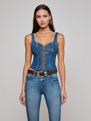 A woman wears the Nayeli Corset Top by L'AGENCE Sale—a sleeveless stretch denim piece with front lace-up detail—paired with blue jeans and a vegan suede brown belt, posing against a plain light background.