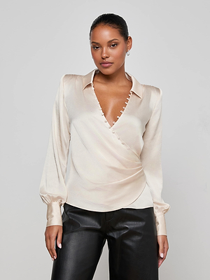 A woman modeling the L'AGENCE Bernice Satin Wrap Blouse in champagne with long sleeves and black leather pants stands against a plain light background, confidently gazing at the camera with romantic elegance.