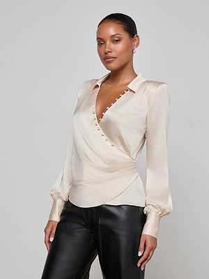 A woman wears the L'AGENCE Bernice Satin Wrap Blouse in cream, styled with black leather pants, standing against a plain light gray background.