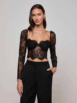 A woman with straight brown hair poses against a plain backdrop, wearing the L'AGENCE Lori Lace Bustier Top—black with floral lace, long sleeves, and a sweetheart neckline—paired with high-waisted black trousers for an elegant evening look.