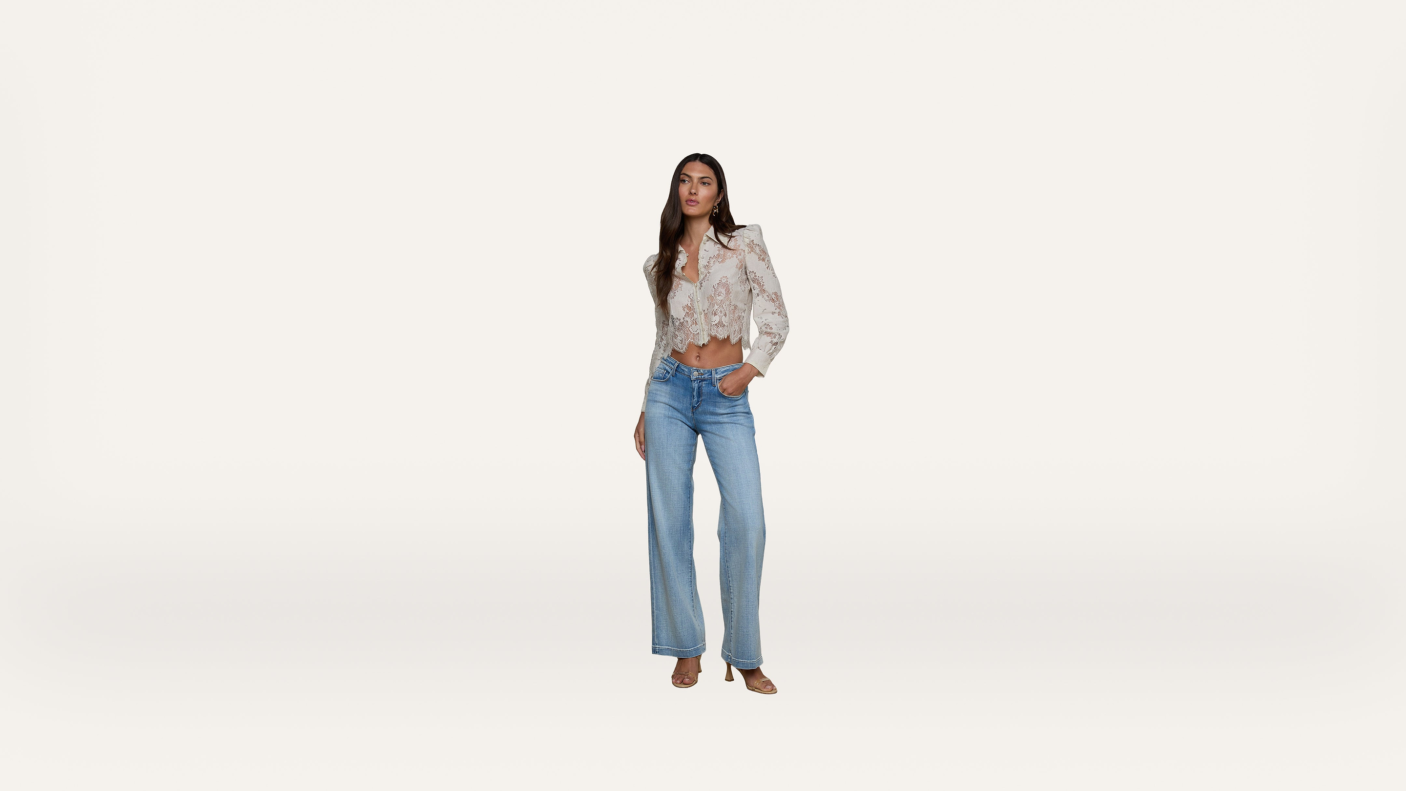 A woman stands against a plain white background, wearing a light floral blouse with puffed shoulders, tucked into high-waisted wide-leg blue jeans and open-toe shoes. She has one hand in her pocket and looks forward.