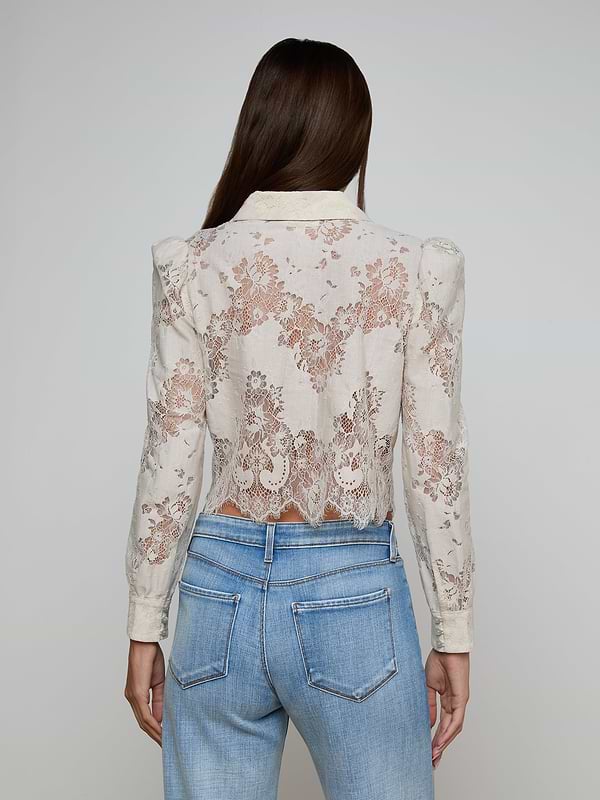 A woman with long brown hair is seen from behind wearing the Anita Lace Blouse, a sheer white floral lace long-sleeve top, paired with light blue jeans that feature high-waisted back pockets. The delicate pattern of the blouse is clearly visible.