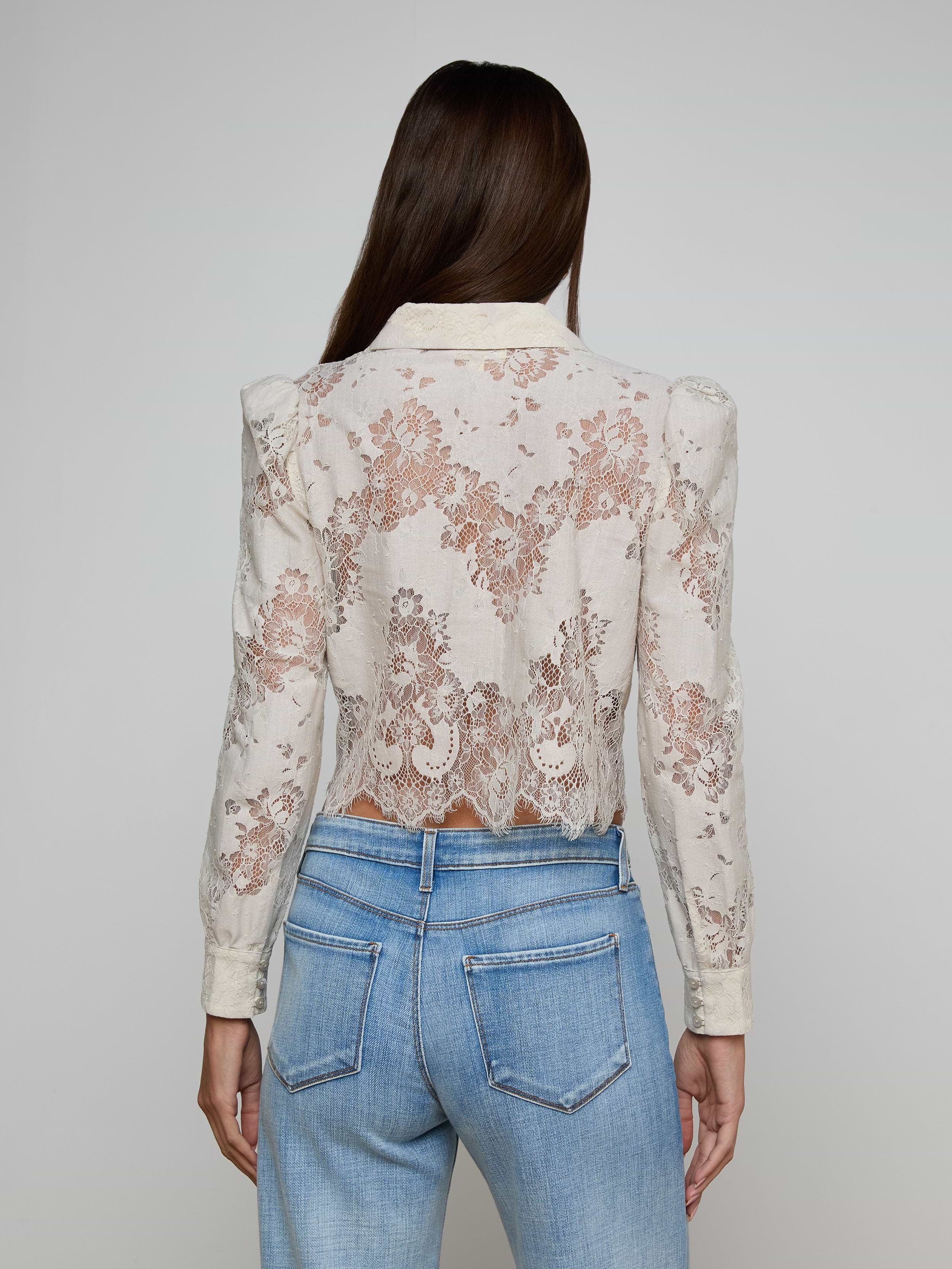 A woman with long brown hair is seen from behind wearing the Anita Lace Blouse, a sheer white floral lace long-sleeve top, paired with light blue jeans that feature high-waisted back pockets. The delicate pattern of the blouse is clearly visible.