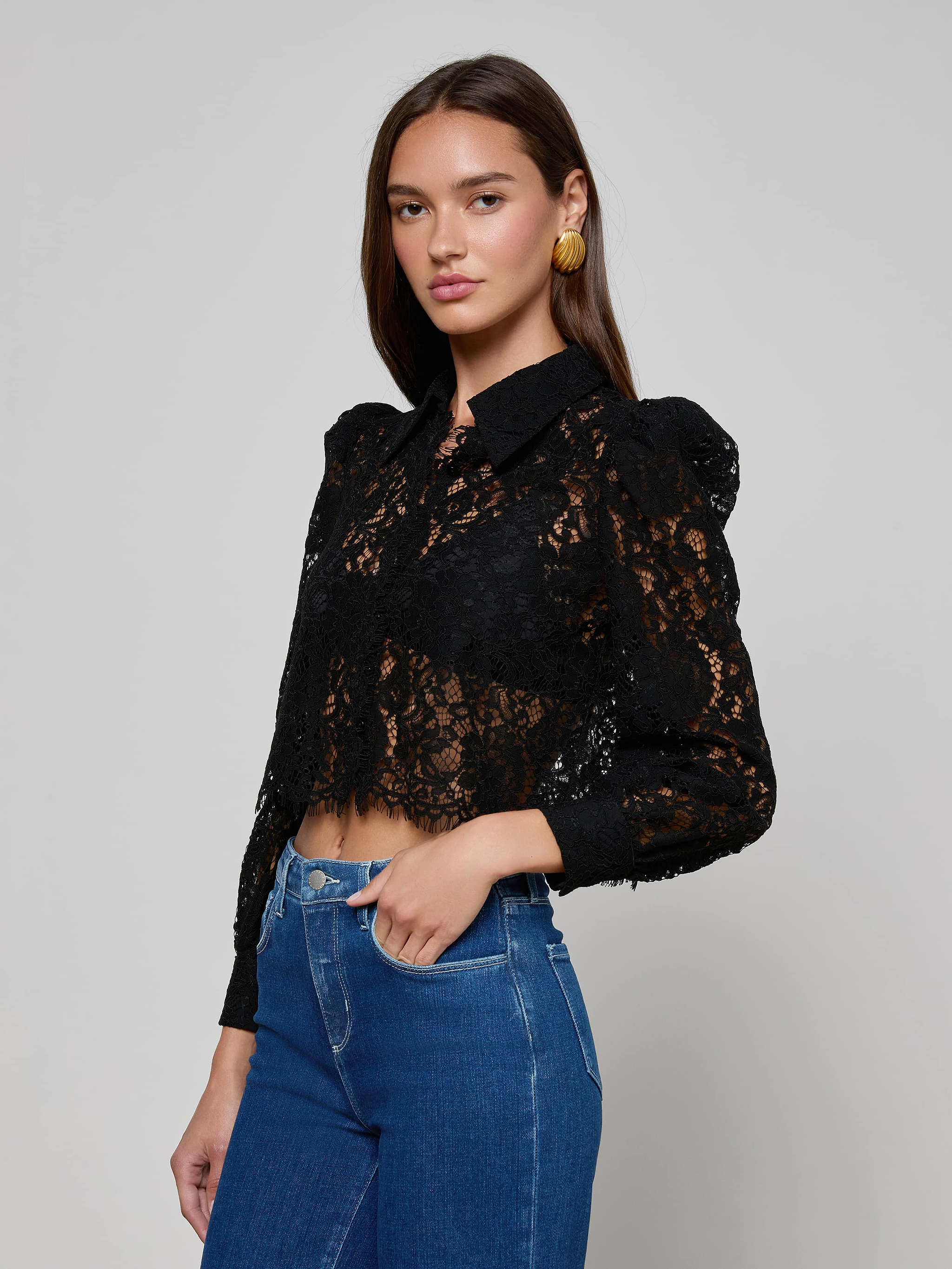 A woman wearing the Anita Lace Blouse—a black floral lace top with puffed shoulders—paired with blue high-waisted jeans, stands against a plain light background.