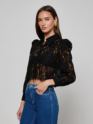 A woman with long brown hair and gold earrings models the L'AGENCE Anita Lace Blouse—black floral lace with puffy shoulders and a scalloped hem—paired with high-waisted blue jeans against a plain background.