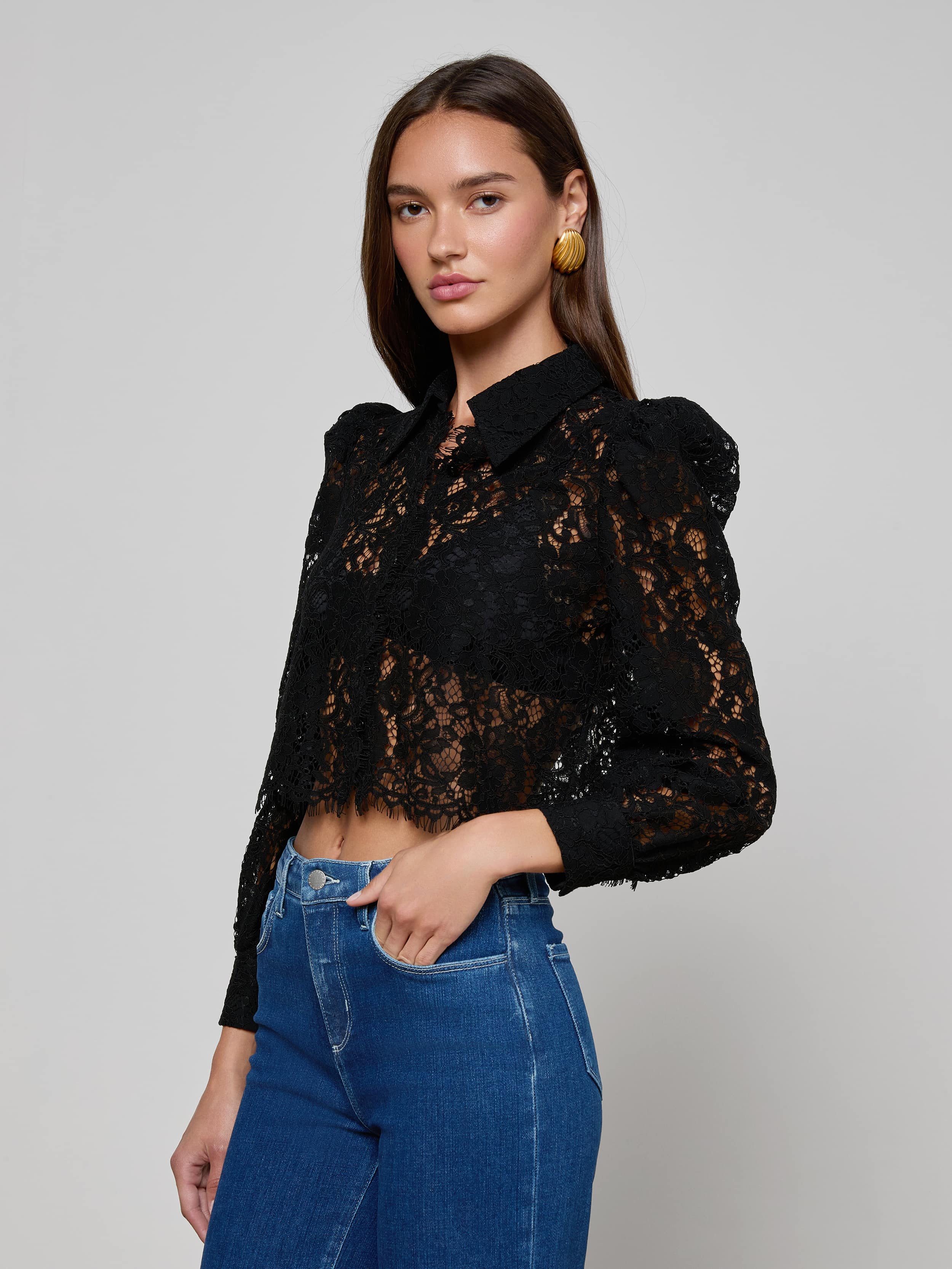 A woman wearing the Anita Lace Blouse—a black floral lace top with puffed shoulders—paired with blue high-waisted jeans, stands against a plain light background.