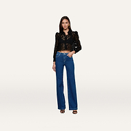 A woman stands facing forward, wearing the Anita Lace Blouse—a black floral lace top with puffed shoulders and a scalloped hem—paired with high-waisted blue wide-leg jeans and black heels against a plain, light background.