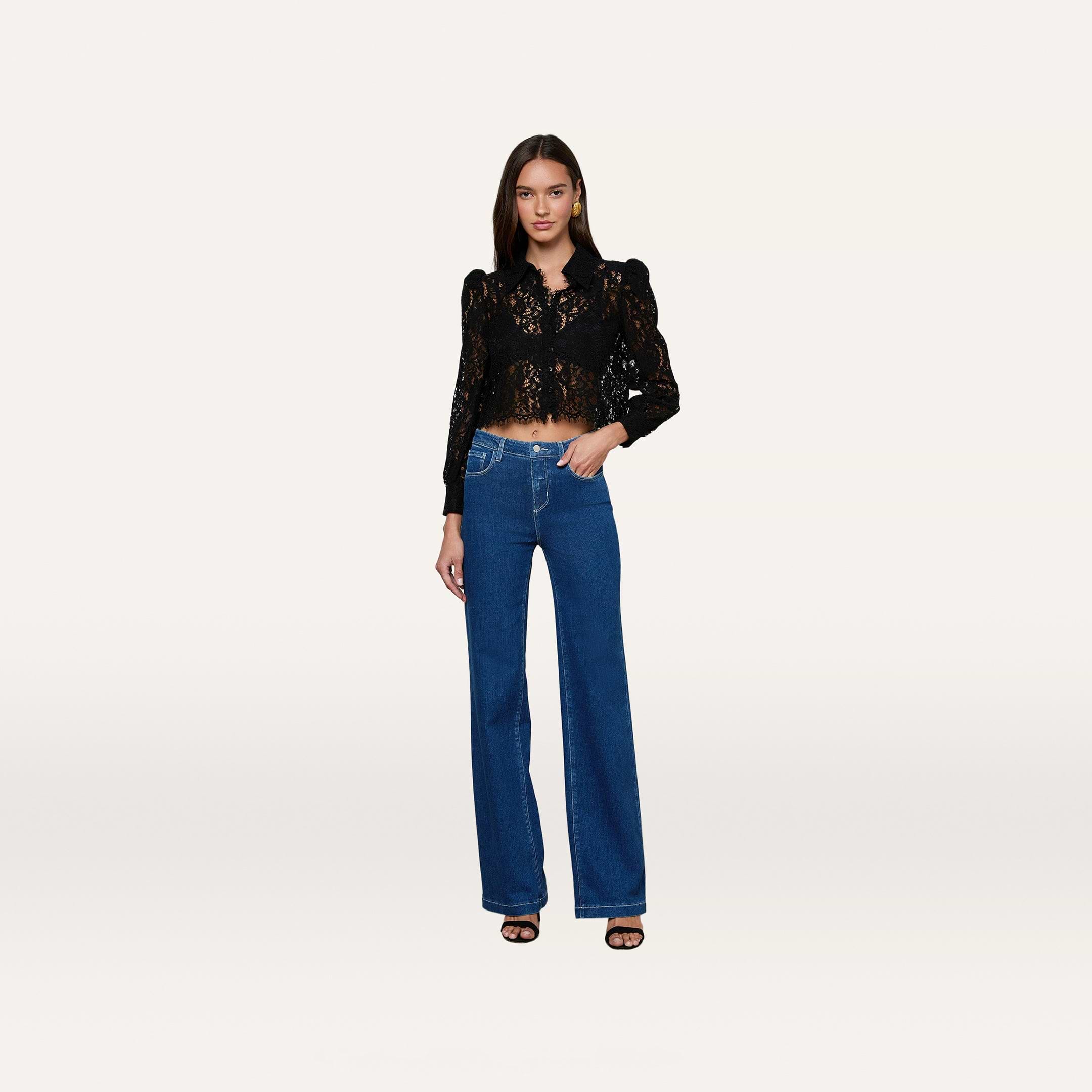 A woman stands facing forward, wearing the Anita Lace Blouse—a black floral lace top with puffed shoulders and a scalloped hem—paired with high-waisted blue wide-leg jeans and black heels against a plain, light background.