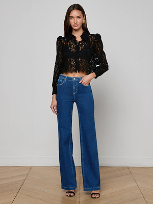 A woman with long brown hair stands indoors against a plain wall, wearing the L'AGENCE Anita Lace Blouse, high-waisted blue jeans, and black heeled sandals, looking at the camera.