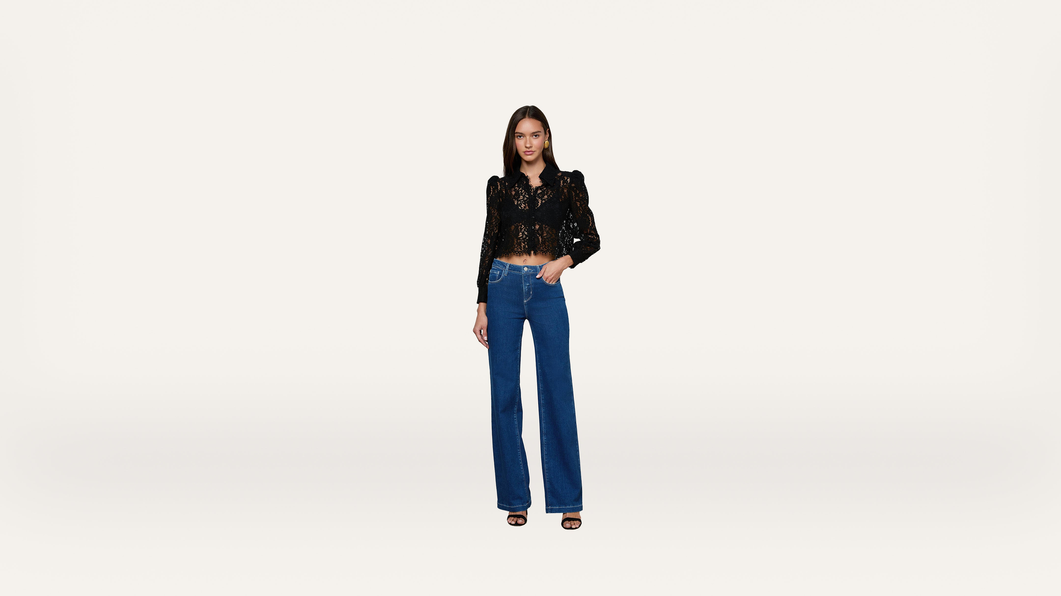 A woman wearing a black lace top, blue wide-leg jeans, and black heeled sandals stands against a plain white background.