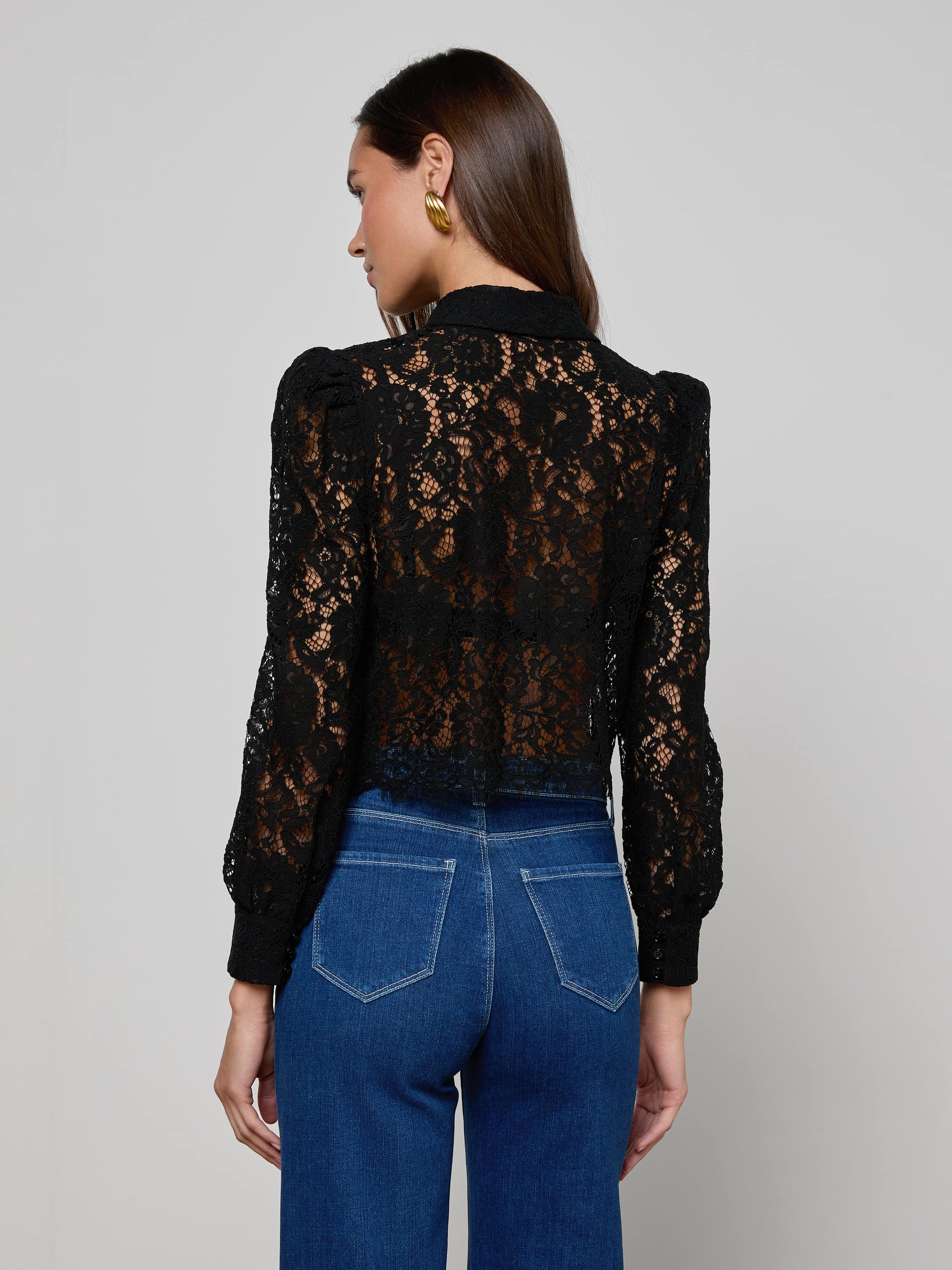 A woman with long brown hair is seen from the back against a plain light background wearing the Anita Lace Blouse, which features floral lace, puffed shoulders, and long sleeves. She pairs it with high-waisted blue jeans.