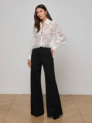A woman poses indoors on a light wooden floor against a white wall, wearing the Anita Lace Blouse—sheer white with floral lace and long sleeves—paired with black wide-leg trousers.