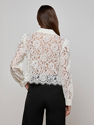 A person with long brown hair is seen from the back, wearing the Anita Lace Blouse—a sheer white floral lace top with long sleeves, an intricate pattern, delicate scalloped hem—and black pants.