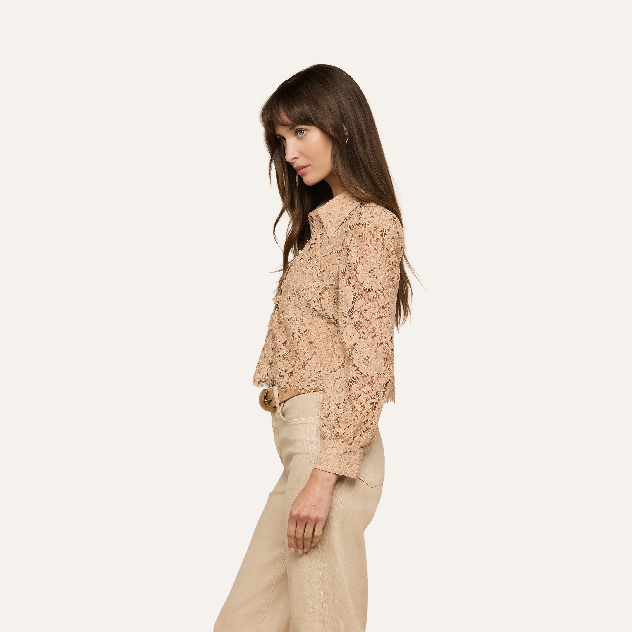 A woman in profile wearing the Anita Lace Blouse, featuring beige floral lace with a scalloped hem, paired with light beige pants, stands against a plain light background.
