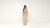 A woman with long hair wears sunglasses, a beige blouse, and matching high-waisted pants, standing against a plain white background.