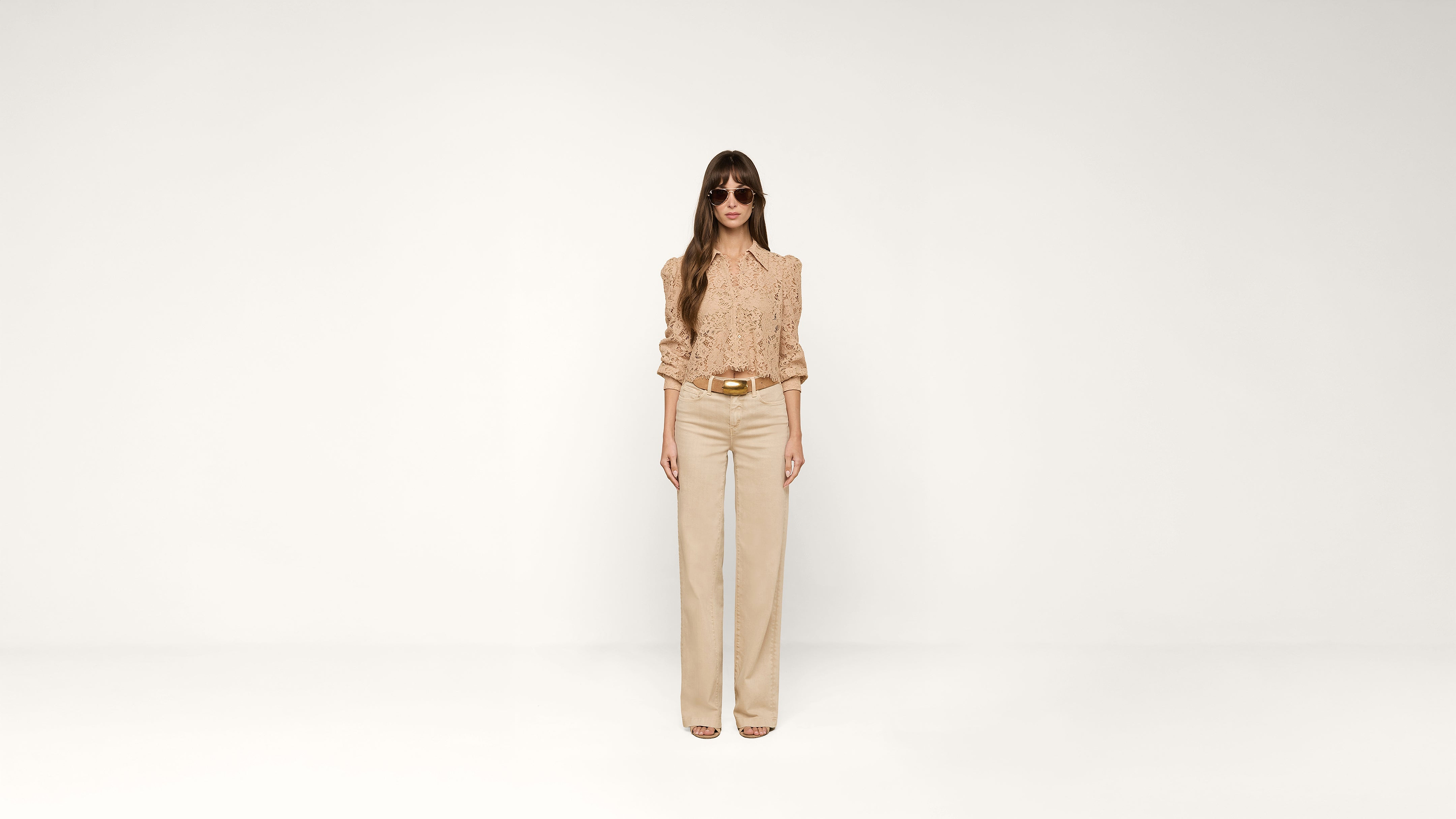 A woman with long hair wears sunglasses, a beige blouse, and matching high-waisted pants, standing against a plain white background.