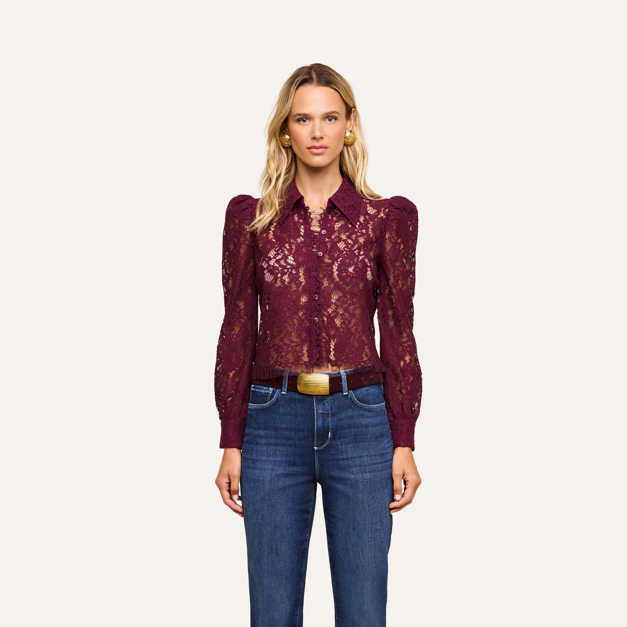 A woman with long blonde hair and a neutral expression stands against a plain background, wearing the Anita Lace Blouse in sheer burgundy floral lace with long sleeves, paired with blue jeans, a belt, and gold earrings.