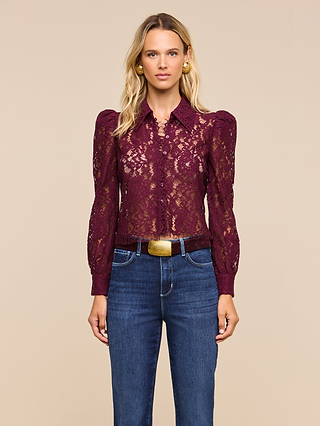 A woman stands against a beige background, wearing the Anita Lace Blouse—a sheer, long-sleeved floral lace top with a collar—paired with dark blue high-waisted jeans. Her loose blonde hair and gold hoop earrings enhance the romantic look of the blouse.