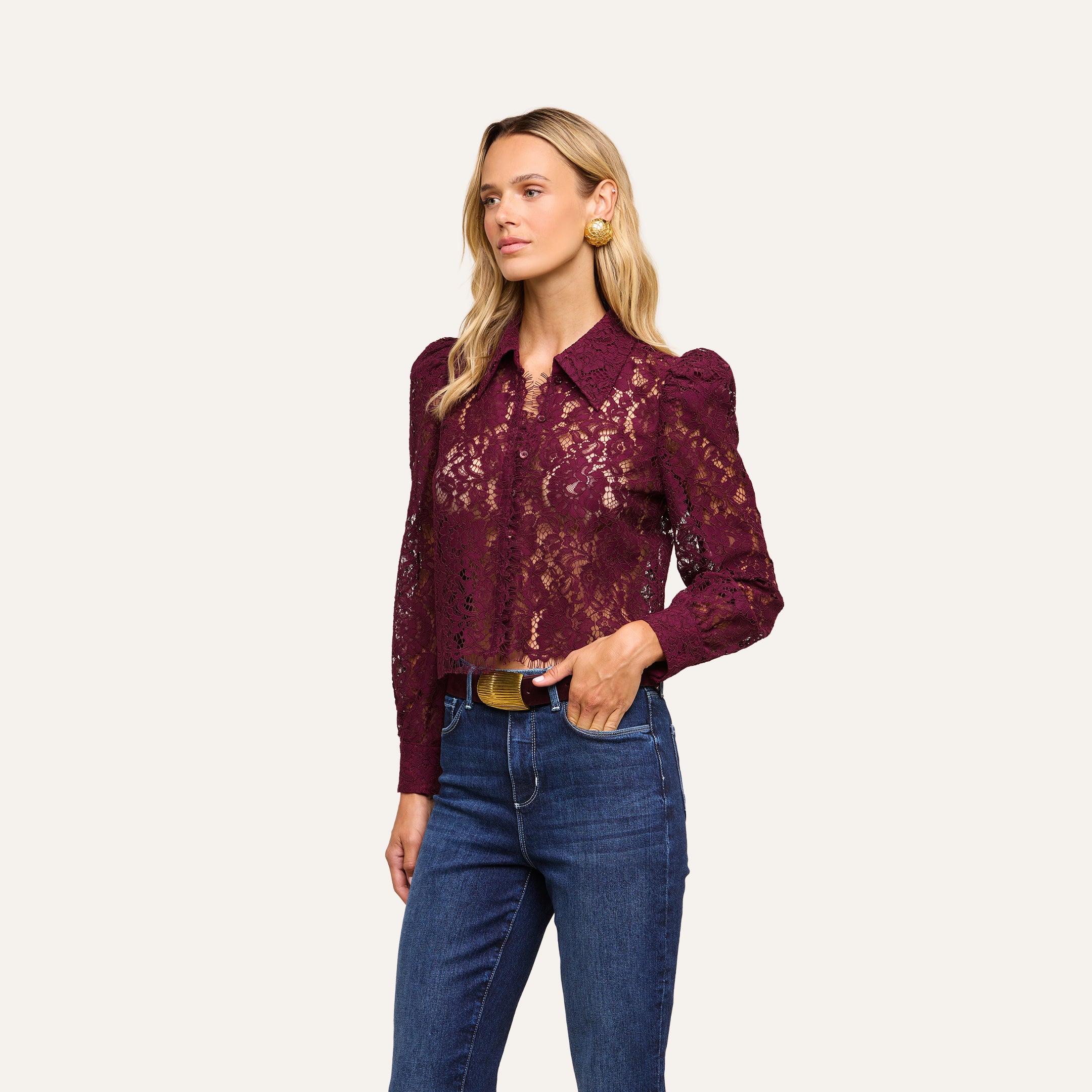 A woman wearing the Anita Lace Blouse—a sheer, burgundy floral lace long-sleeve top—paired with blue jeans, a black belt with a gold buckle, and gold earrings, stands against a plain light background.