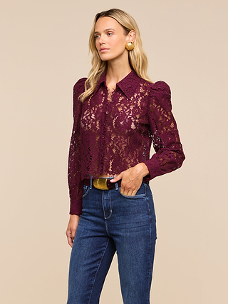 Wearing the Anita Lace Blouse in burgundy with a floral lace design and long sleeves, paired with blue jeans and gold earrings, a woman stands against a beige background, glancing to the side.
