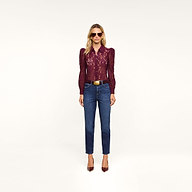 A woman wearing sunglasses, the romantic burgundy Anita Lace Blouse with puffed sleeves, blue jeans, a belt, and burgundy heels stands against a plain white background.