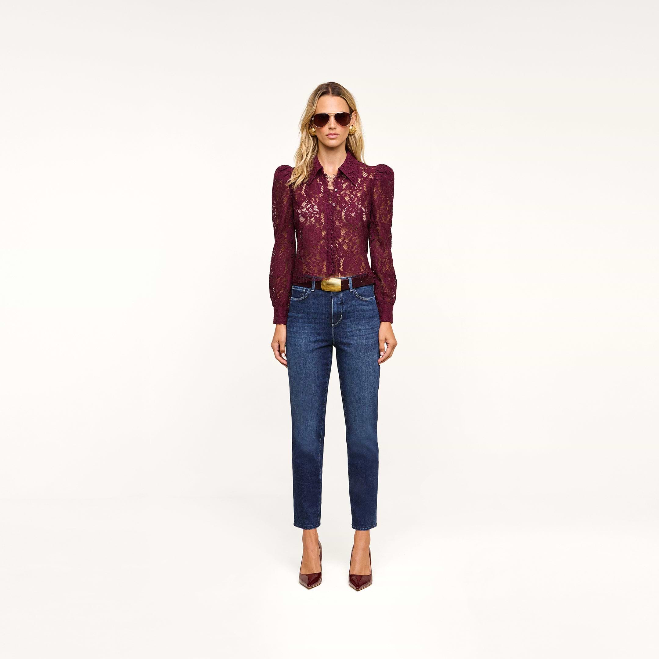 A woman wearing sunglasses, the romantic burgundy Anita Lace Blouse with puffed sleeves, blue jeans, a belt, and burgundy heels stands against a plain white background.