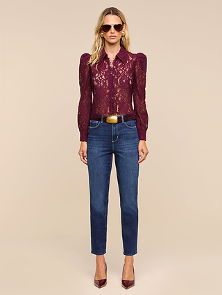 A woman wearing sunglasses, the Anita Lace Blouse—a romantic burgundy floral lace top with long puffed sleeves—paired with blue jeans, a brown belt, and burgundy heels, stands against a plain beige background.