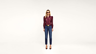 A woman wearing sunglasses, a maroon blouse, blue high-waisted jeans, and maroon heels stands against a plain white background, facing forward.