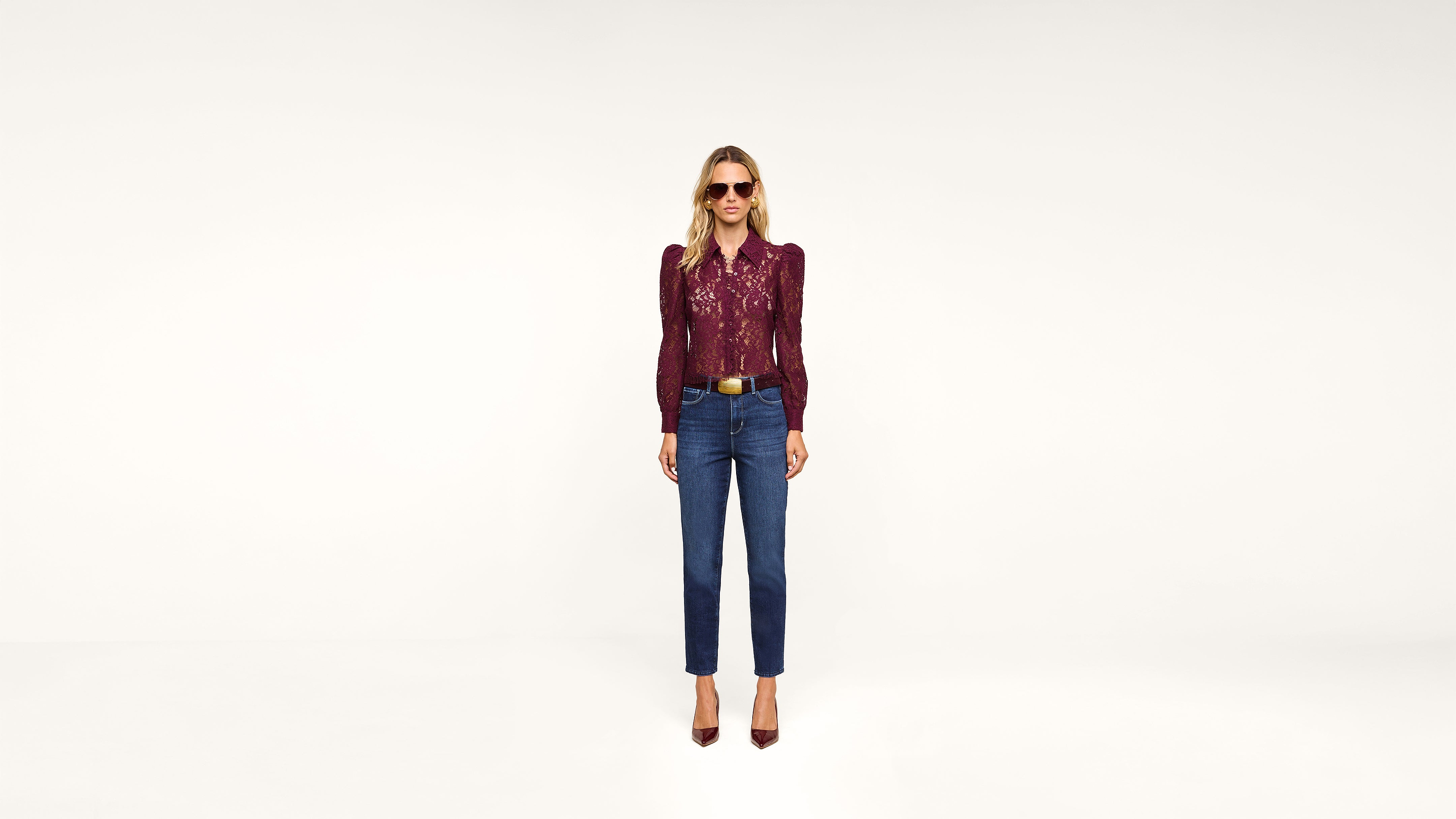 A woman wearing sunglasses, a maroon blouse, blue high-waisted jeans, and maroon heels stands against a plain white background, facing forward.