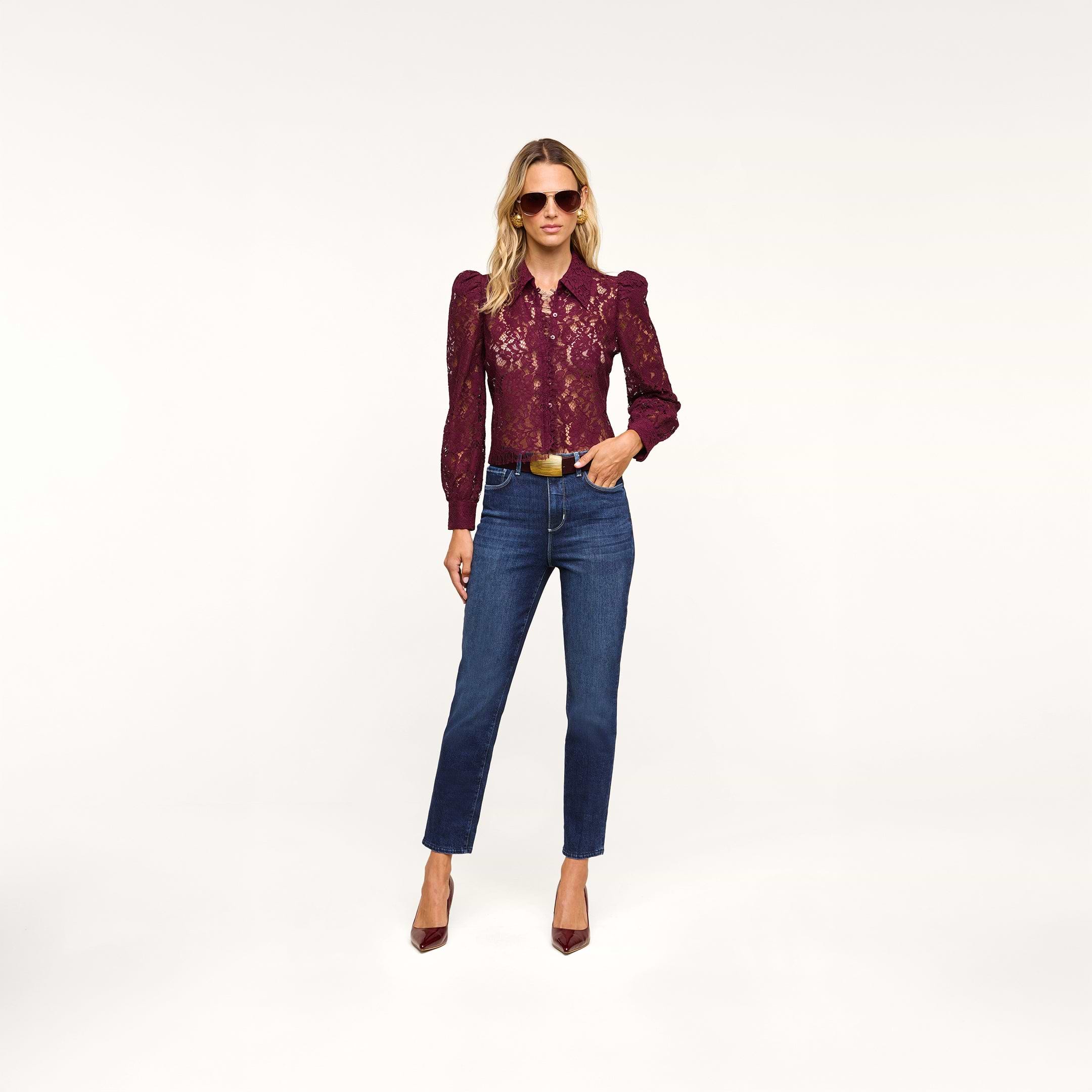 A woman wearing sunglasses, the Anita Lace Blouse in burgundy, blue jeans, a brown belt, and burgundy heels stands against a plain white background.