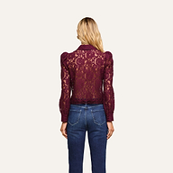 A woman stands facing away, wearing the Anita Lace Blouse—a sheer, long-sleeved, burgundy floral lace top—paired with blue jeans. Her blonde hair is loose, and the background is plain and off-white.