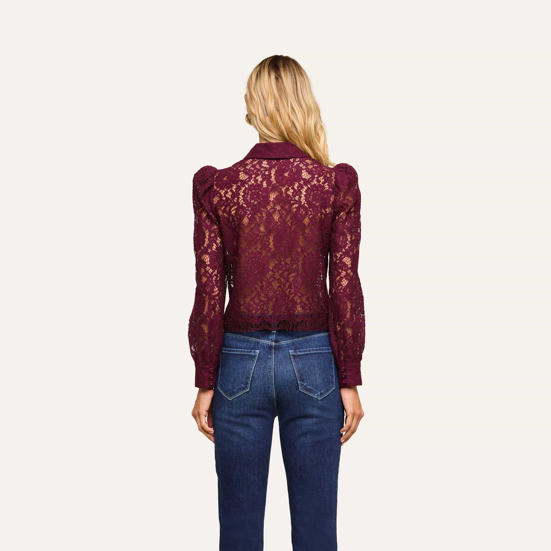 A woman stands facing away, wearing the Anita Lace Blouse—a sheer, long-sleeved, burgundy floral lace top—paired with blue jeans. Her blonde hair is loose, and the background is plain and off-white.
