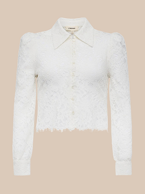 The Anita Lace Blouse is a romantic white long-sleeve top featuring floral lace, a collared button-down front, puffed shoulders, and a slightly frayed hem, shown against a beige background.