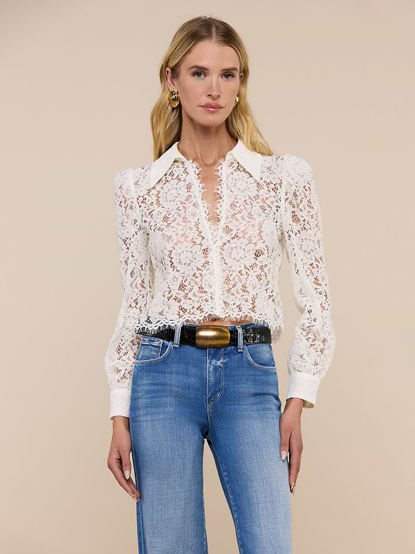 Wearing the Anita Lace Blouse, a woman showcases its romantic sheer white floral lace and long sleeves, pairing it with blue jeans, a black belt with a gold buckle, and gold earrings as she stands against a plain light background.