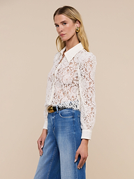 Wearing the Anita Lace Blouse—a romantic sheer white floral lace blouse with a collar and long sleeves—paired with blue high-waisted jeans and gold earrings, a woman stands against a plain light background.