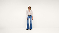 A woman wearing sunglasses, a patterned long-sleeve blouse, wide-leg blue jeans, a black belt, and black shoes stands against a plain white background.
