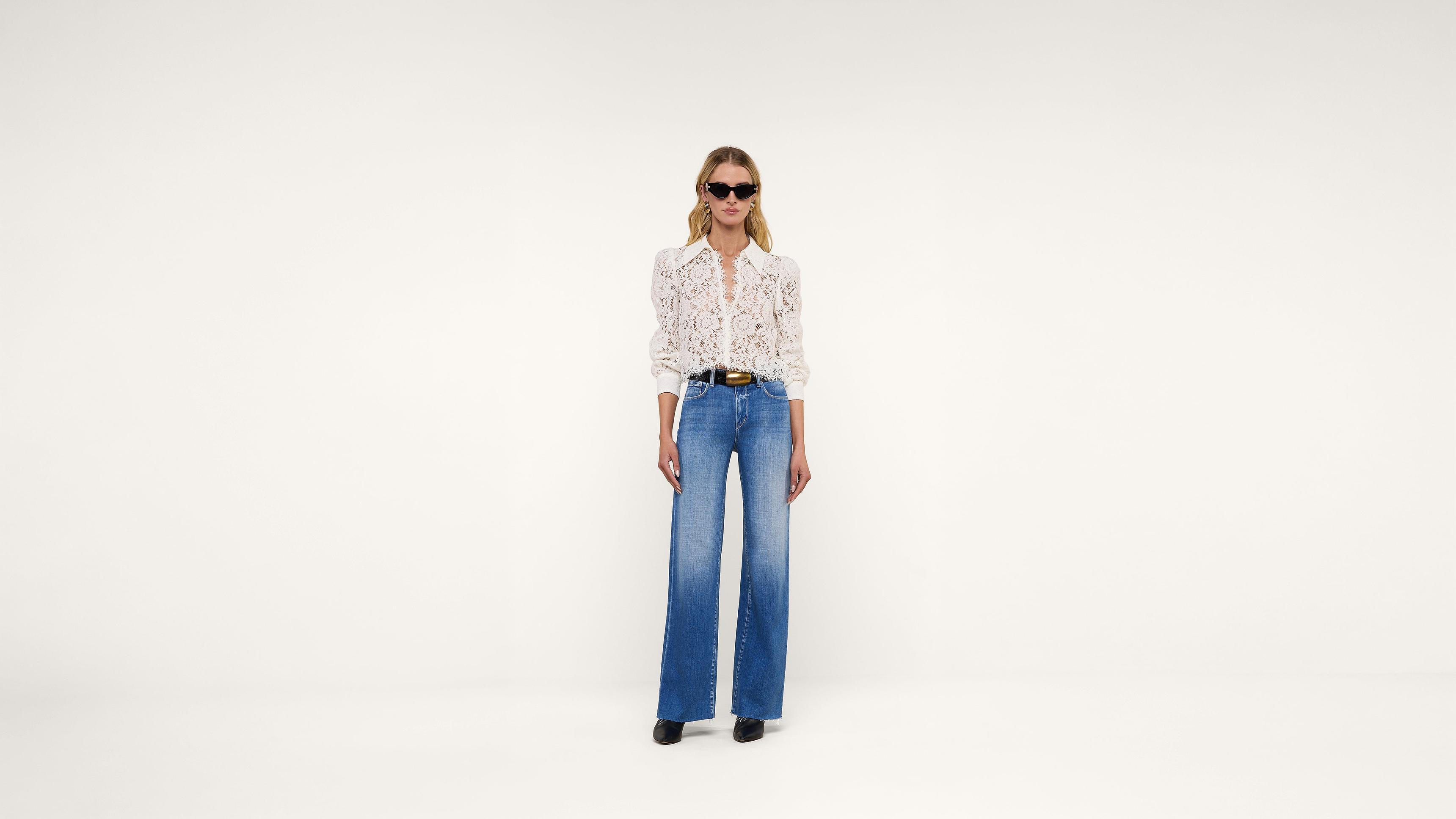 A woman wearing sunglasses, a patterned long-sleeve blouse, wide-leg blue jeans, a black belt, and black shoes stands against a plain white background.