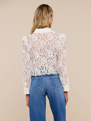 A woman with long blonde hair, seen from behind, wears the Anita Lace Blouse—a sheer white floral lace top with long sleeves—paired with blue high-waisted jeans against a plain beige background.