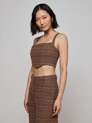 A woman with a bob haircut wears the Tiffany Corset Top by L'AGENCE in brown and red houndstooth, paired with matching high-waisted pants. She stands against a light gray background, smiling softly at the camera.