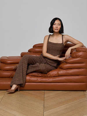 A woman with shoulder-length dark hair sits on a brown leather sofa, wearing the L'AGENCE Tiffany Corset Top and tan heels. The scene features a light gray wall and wooden herringbone floor.
