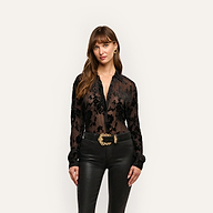 A woman wears a black Laurent Velvet Blouse with a floral devoré pattern, tucked into black pants with a large gold-buckled belt.