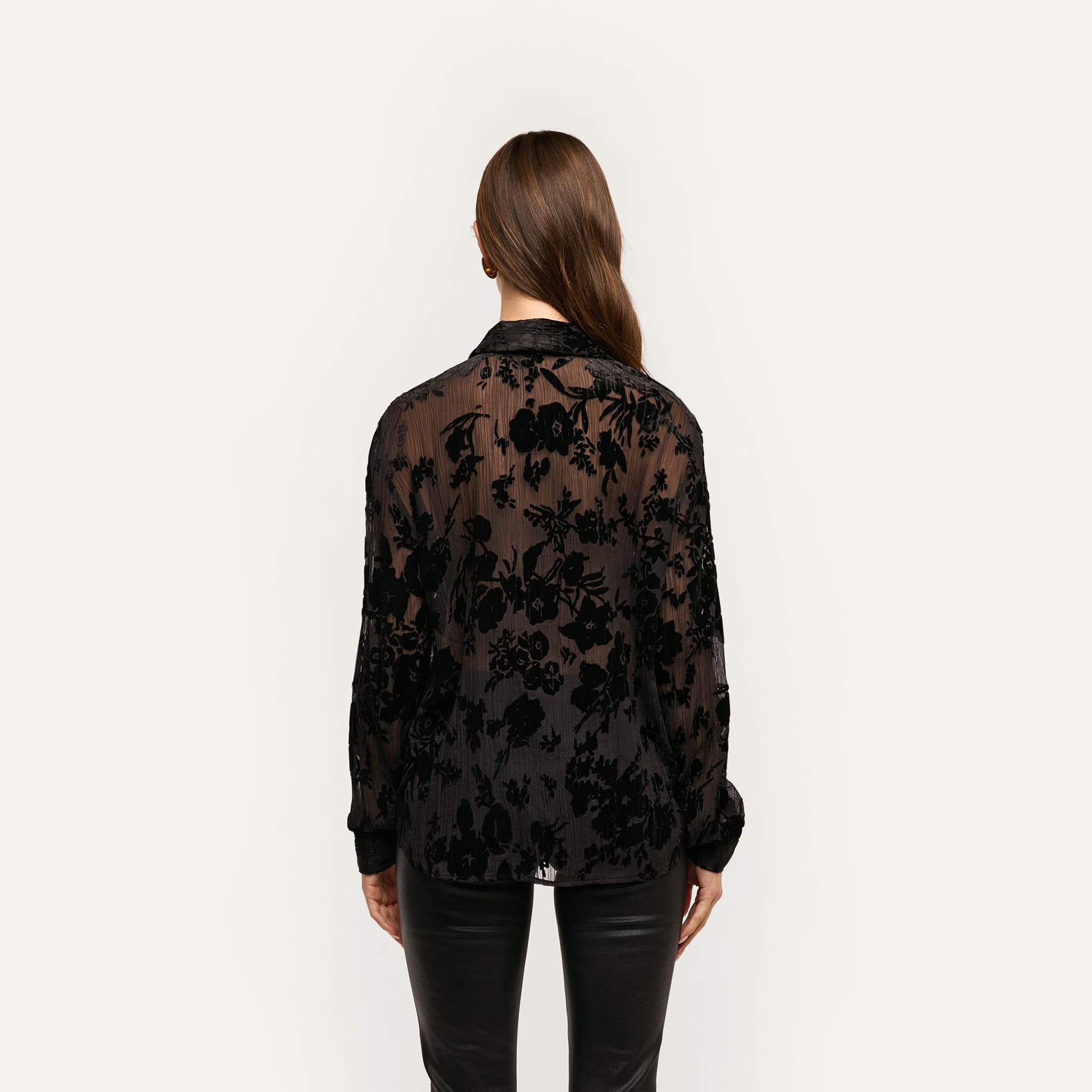 Back view of a person wearing a sheer black Laurent Velvet Blouse with a crinkle velvet floral pattern in a relaxed, button-down fit.