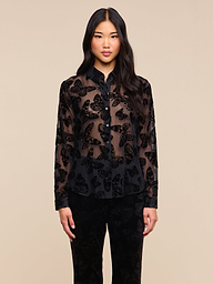 A woman with long black hair stands facing forward against a plain light background, wearing the Laurent Velvet Blouse—featuring a burnout velvet butterfly pattern—paired with black pants.