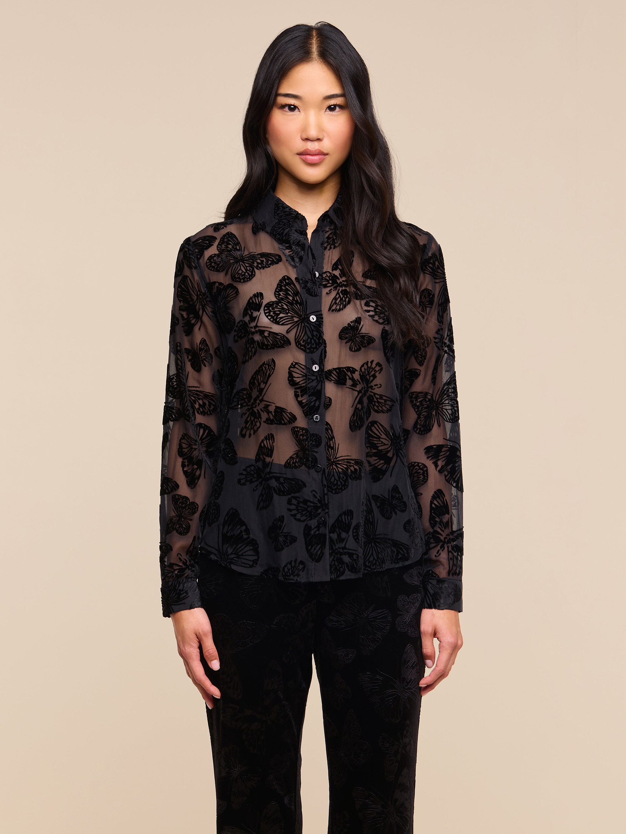 A woman with long black hair stands facing forward against a plain light background, wearing the Laurent Velvet Blouse—featuring a burnout velvet butterfly pattern—paired with black pants.