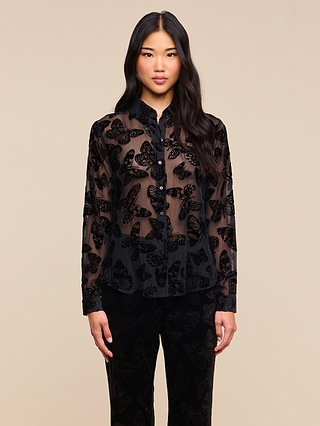 A woman with long dark hair stands against a plain light beige background, facing forward with a neutral expression. She is wearing the Laurent Velvet Blouse—a relaxed fit burnout velvet top featuring a butterfly print—paired with black pants.